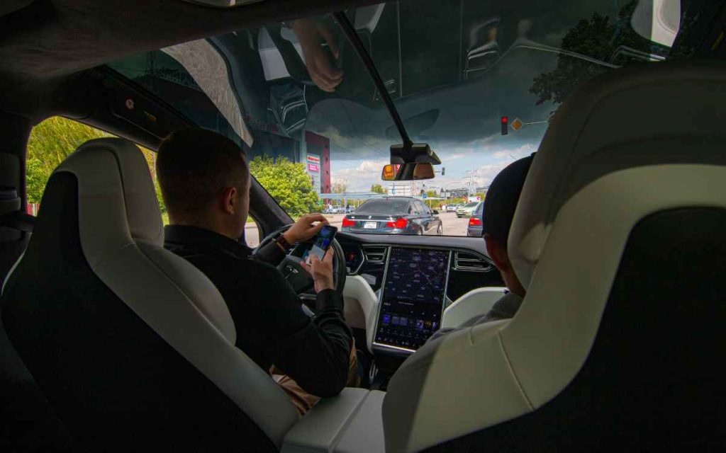 Vemos una imagen de un conductor dentro de un auto usando su teléfono celular, haciendo referencia al accidente del tesla en China.